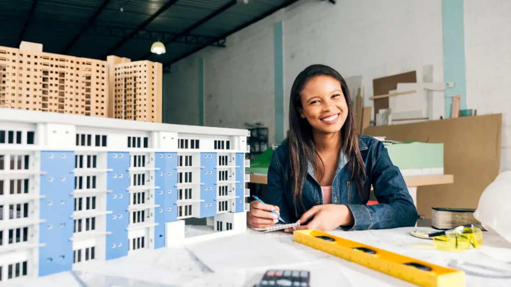 Go-to-market imobiliário:  U imagem mostra uma mulher sentada em uma mesa, olhando para frente e sorrindo. Ela segura um bloco de notas e uma caneta. À sua frente, na mesa, estão maquetes detalhadas de edifícios, uma delas em primeiro plano com fachadas azuis e brancas e outra, de madeira, ao fundo. A mesa está repleta de ferramentas de arquitetura e construção, como uma trena amarela, um nível, uma calculadora e cadernos. 