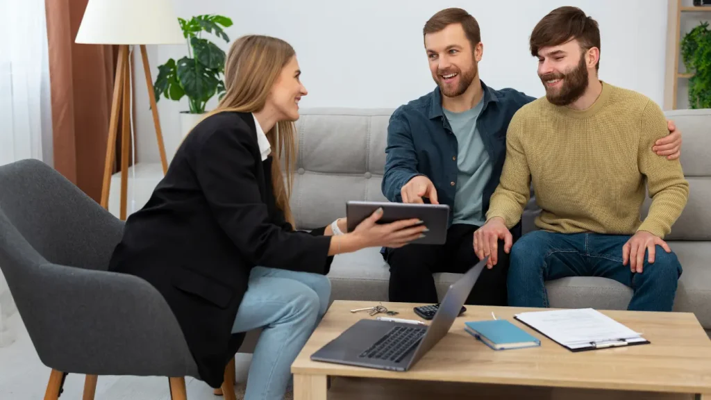 canal de vendas imobiliário: A composição da cena é centrada na interação entre três indivíduos, com uma mulher sentada numa poltrona à esquerda , mostrando um tablet, e o casal de homens acomodado num sofá à direita. A mesa de centro, posicionada entre eles, contém elementos de trabalho como um portátil, papéis, chaves e um computador. Ao fundo da cena há uma planta e uma luminária.