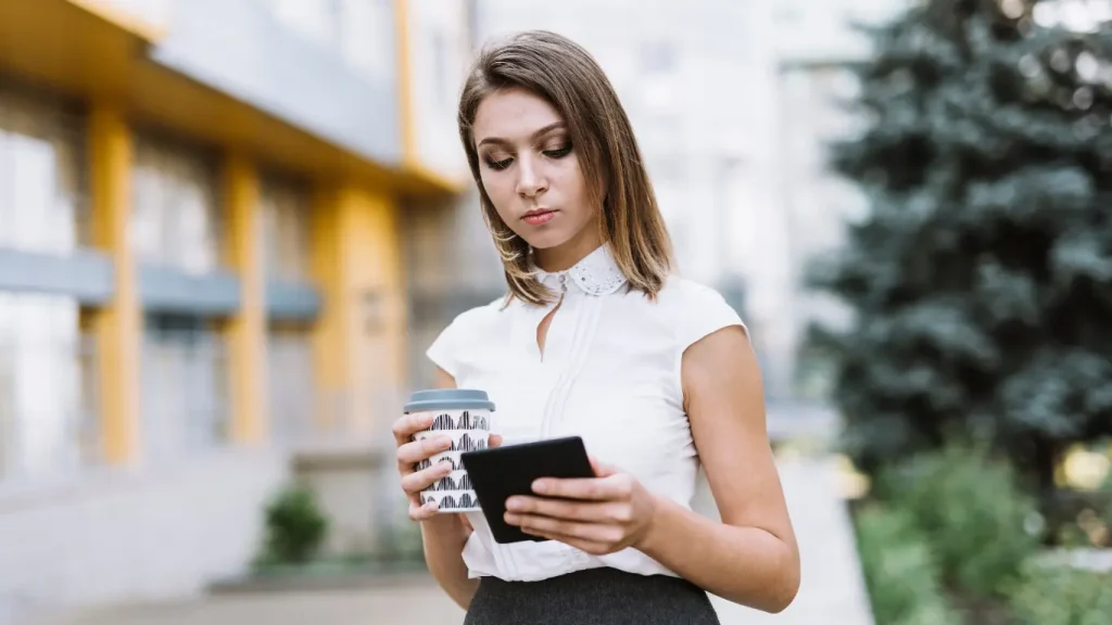 go-to-market imobiliário: Uma mulher parada, posicionada no centro da imagem, com seu olhar voltado para baixo para o tablet em suas mãos. Na outra mão ela segura um copo de café. O fundo da imagem mostra um prédio e uma árvore desfocados.