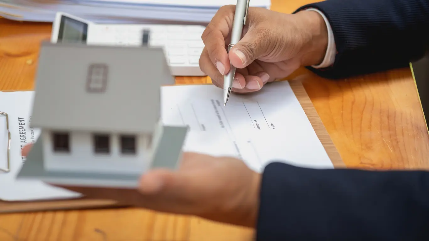 Pessoa assinando um documento relacionado a um imóvel, enquanto segura uma maquete de uma casa sobre uma mesa, representando negociação e contrato imobiliário. pré-lançamento imobiliário