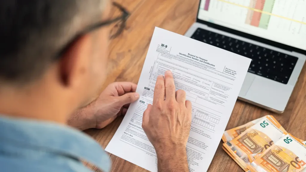 Plantão de vendas imobiliário: Um homem analisa atentamente um formulário financeiro ao lado de um computador portátil e de notas de euro, numa cena que sugere o preenchimento de documentos fiscais ou administrativos.